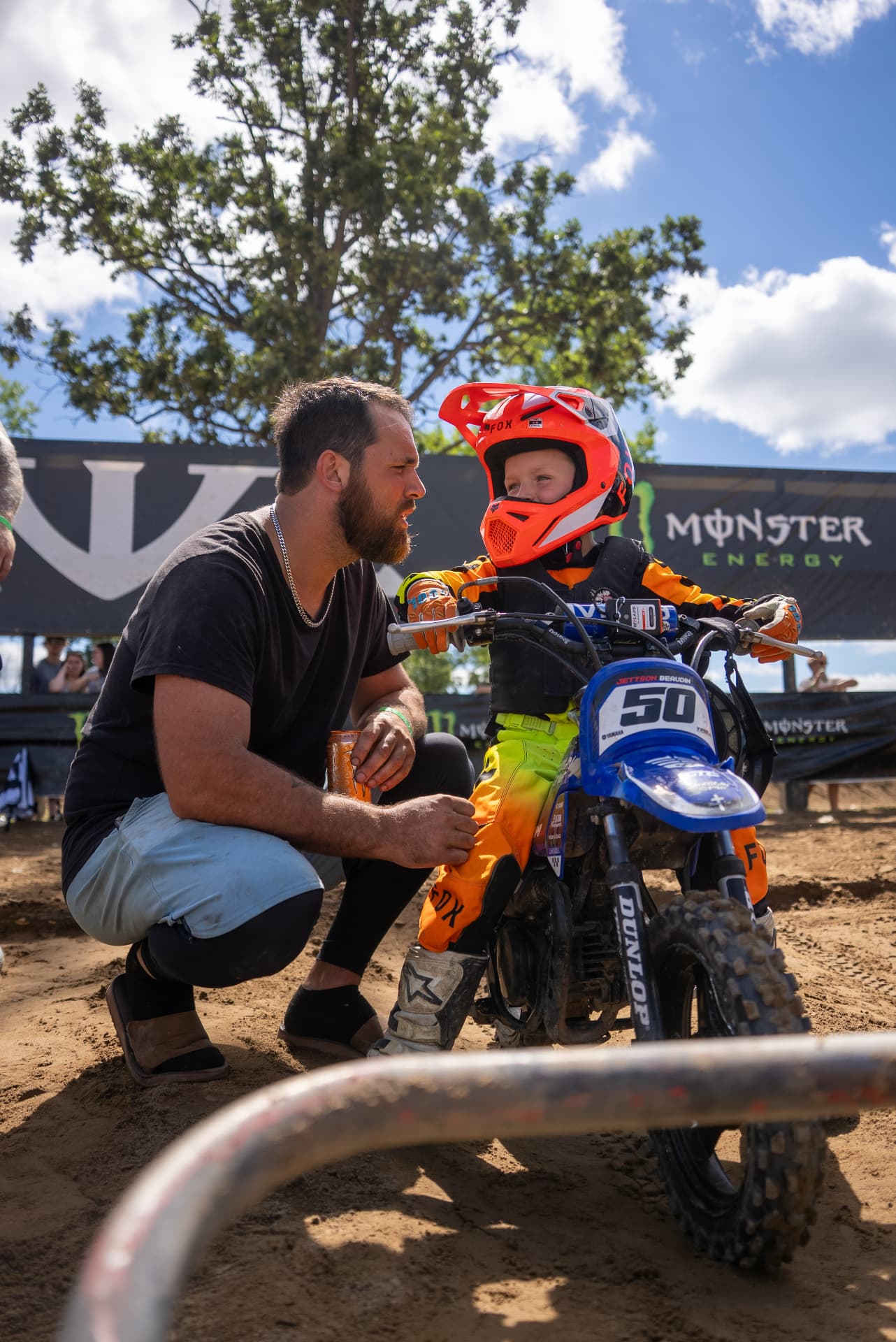 Adult mentor kneeling beside a young amateur motocross racer at the track.