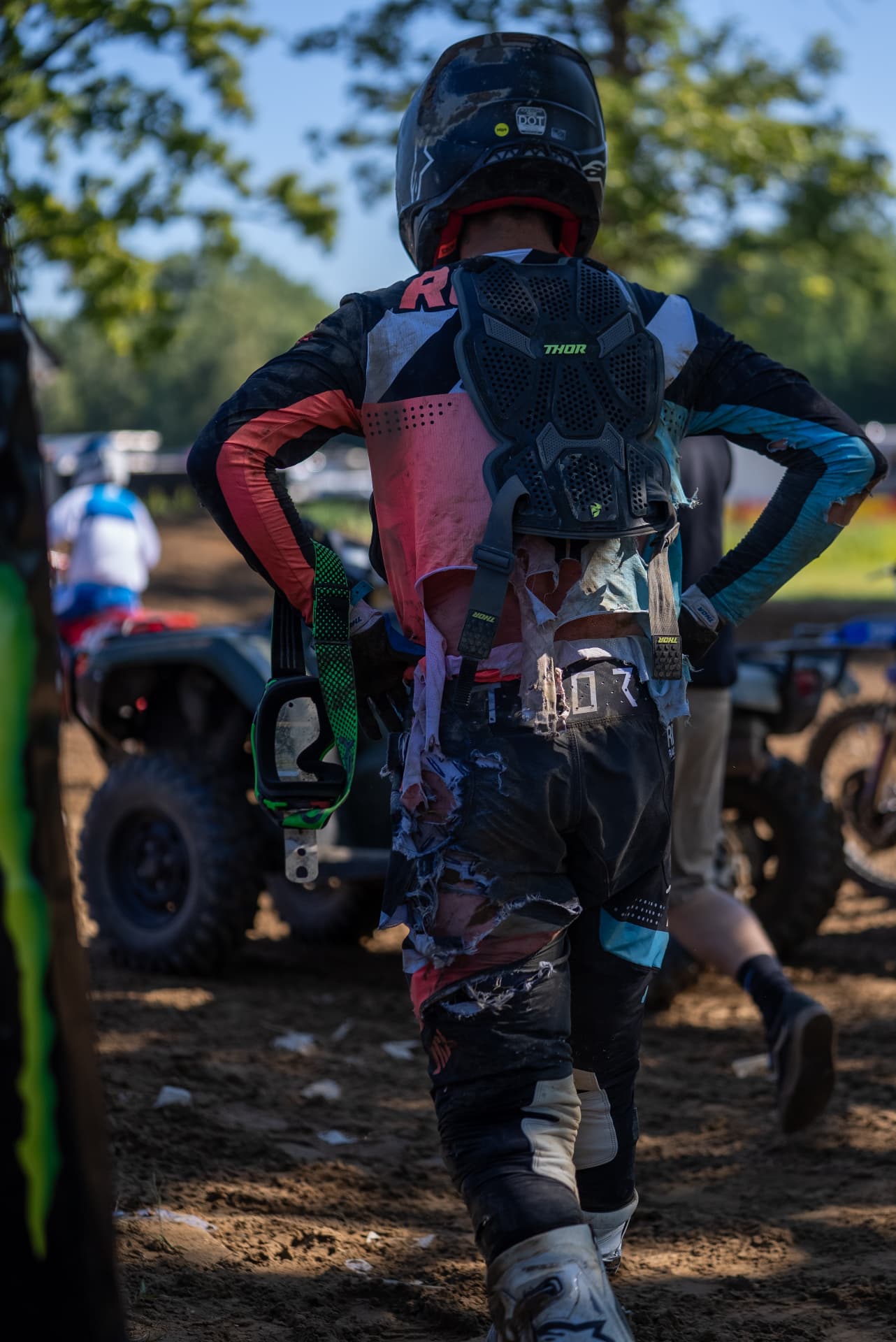 A racer in torn gear walks through the pits, dirt bikes and race day activity in the background.